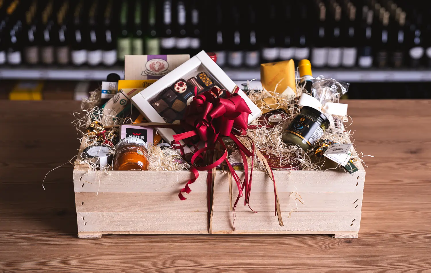 Auf dem Bild sieht man einen Geschenkkorb aus Holz. Darin befinden sich verschiedene Delikatessen und es ist mit einer roten Schleife verziert.