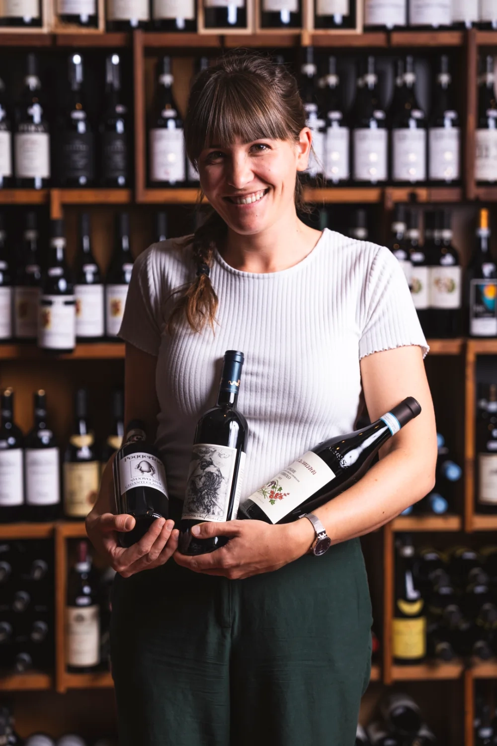 Sabine Hofer steht vor einem Weinregal und hält drei Weinflaschen in den Armen.