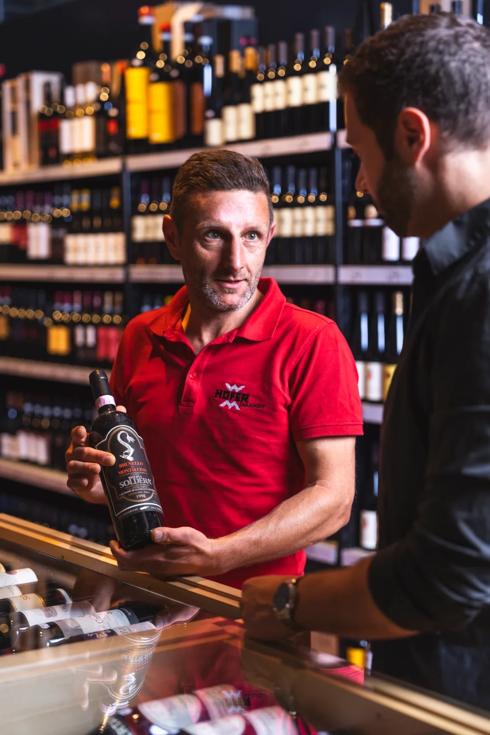 Ein Angestellter des Hofer Market hält eine Flasche Wein in der Hand und berät einen Kunden.