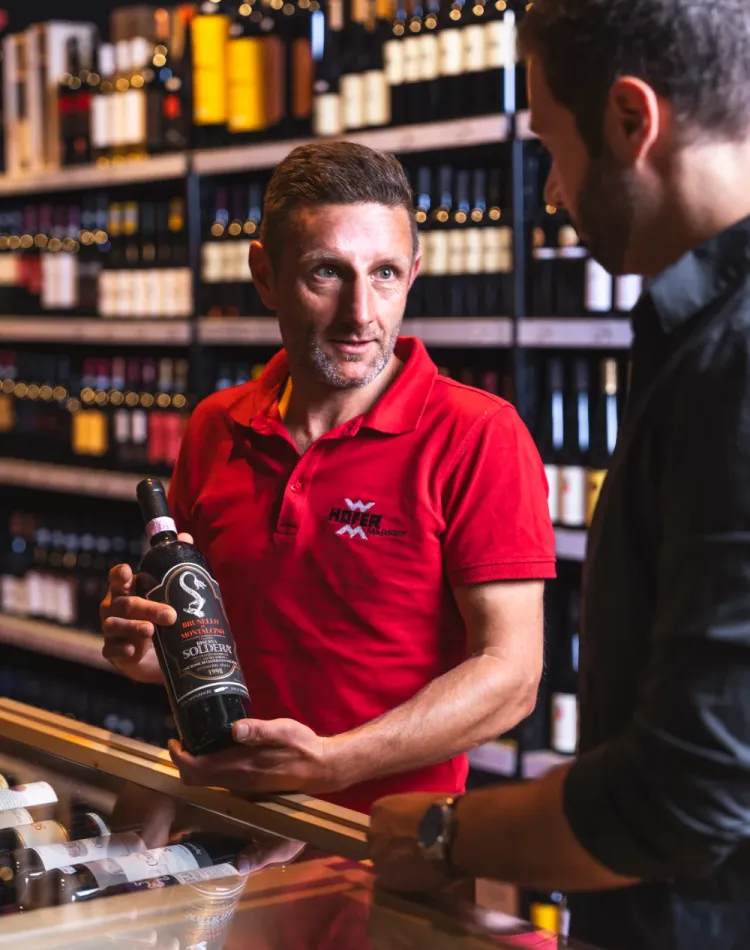 Ein Angestellter des Hofer Market hält eine Flasche Wein in der Hand und berät einen Kunden.
