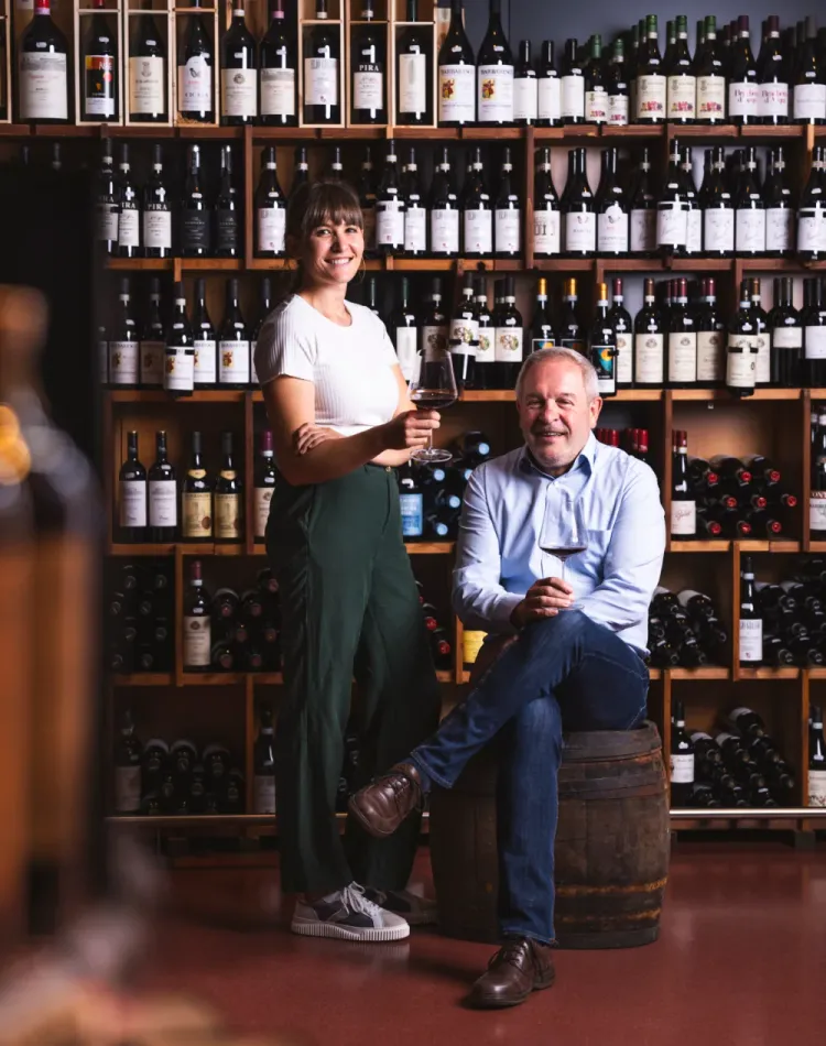Sabine Hofer und Toni Hofer sind vor einem Weinregal und haben jeweils ein Glas Rotwein in der Hand.