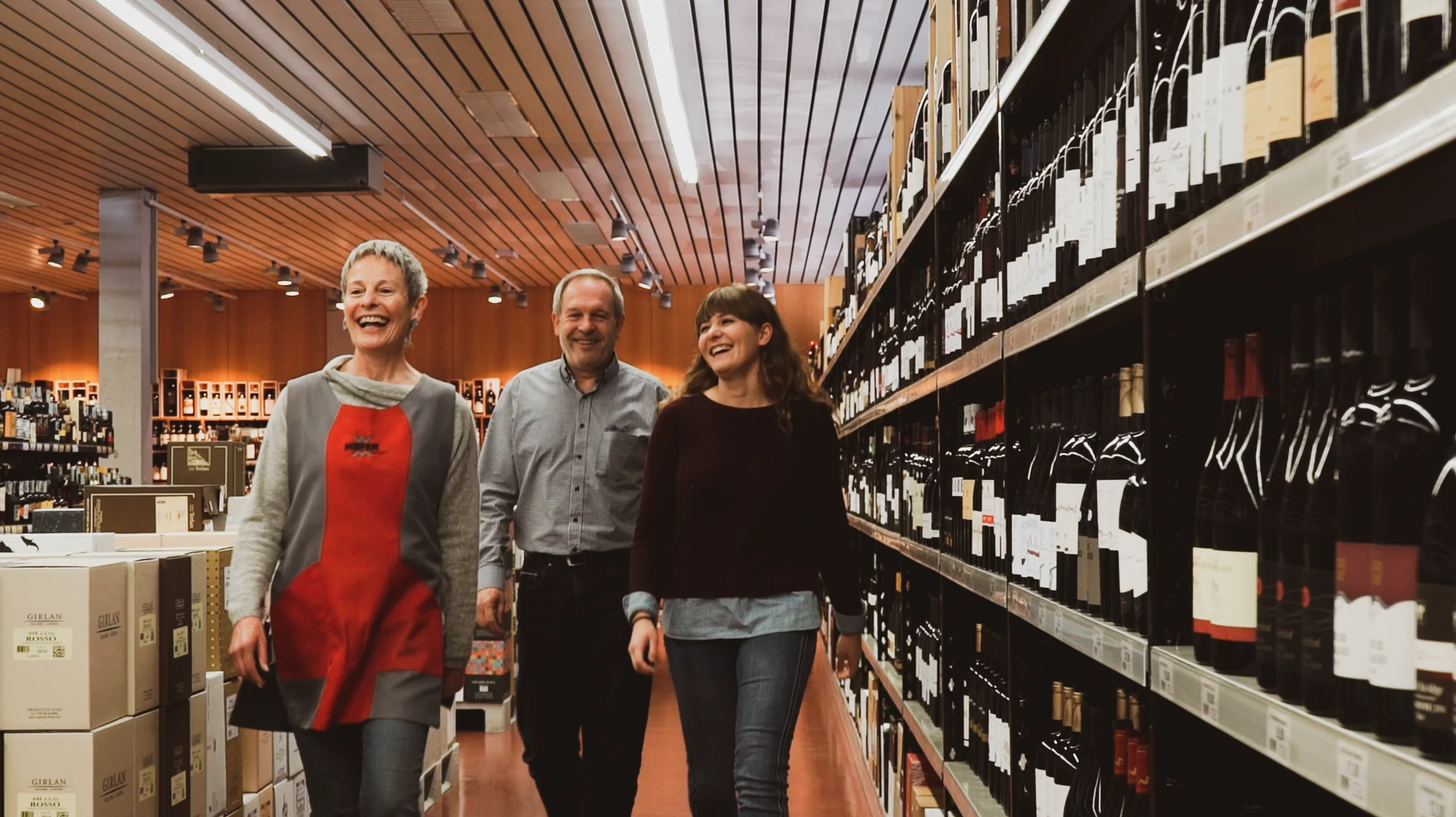 Veronika, Toni und Sabine Hofer gehen an einem Weinregal vorbei und lachen.