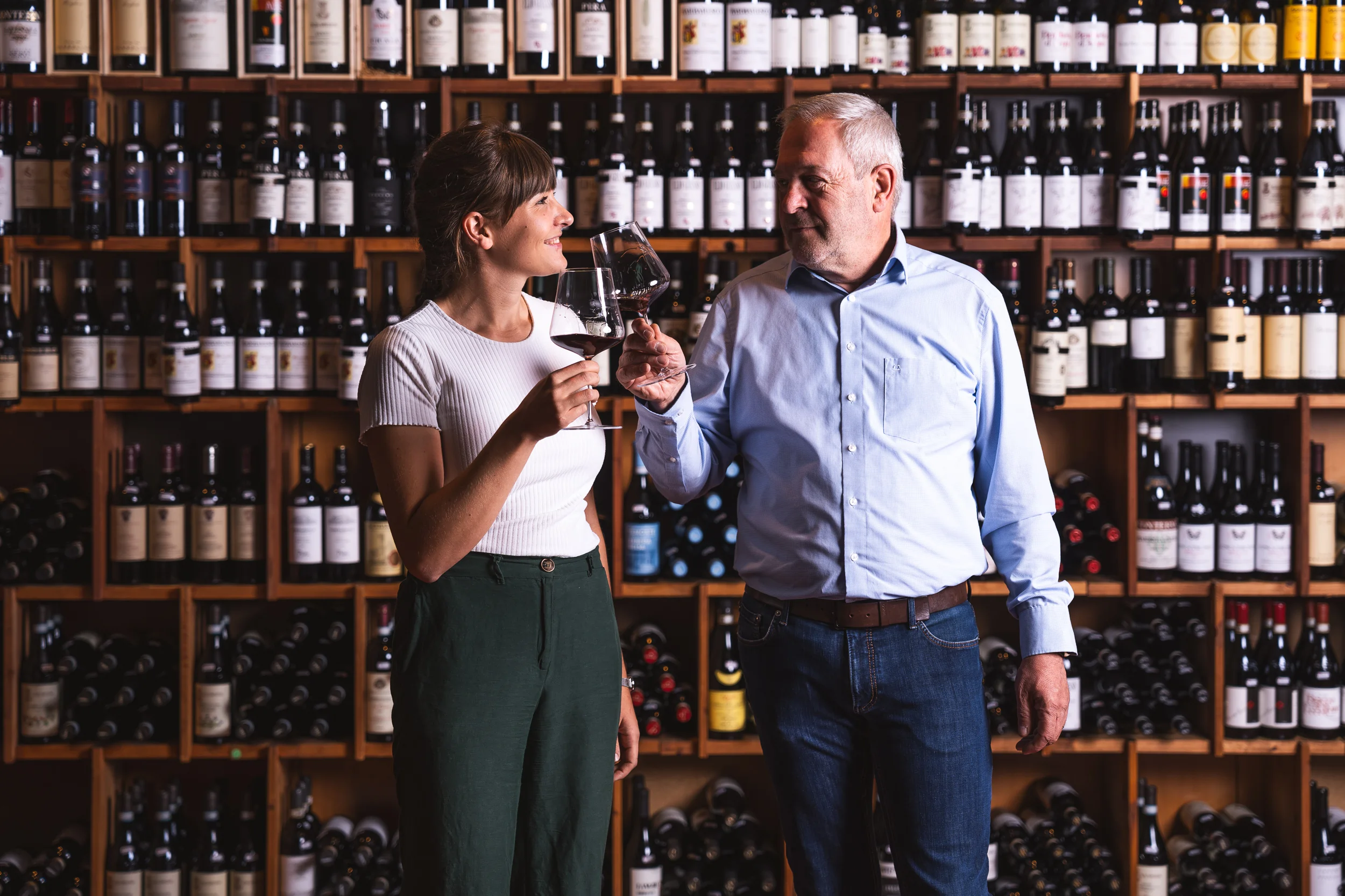 Sabine und Toni Hofer stoßen mit einem Rotweinglas an, dahinter befindet sich ein Rotweinregal.