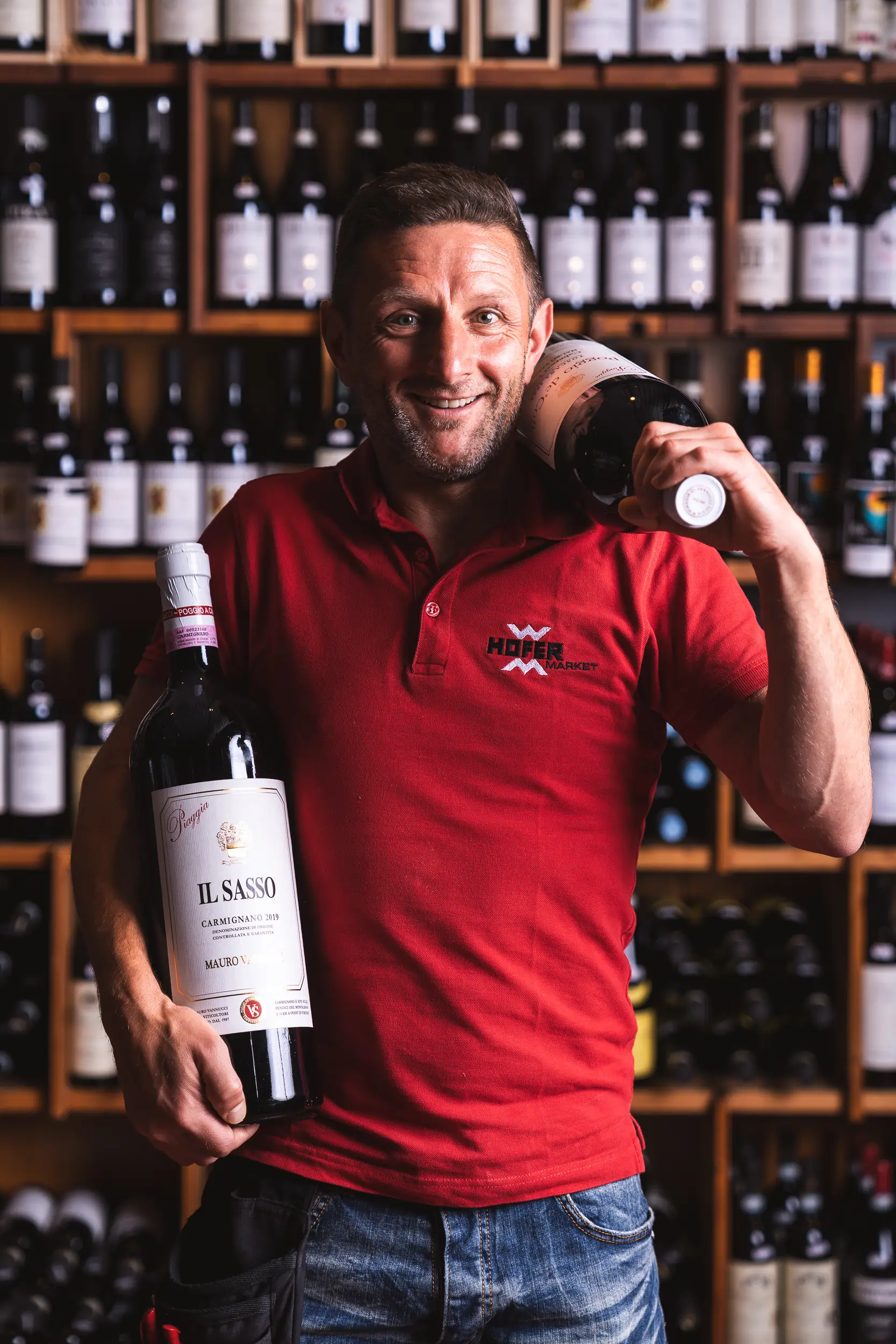 Ein Mitarbeiter des Hofer Market trägt ein rotes T-Shirt und hält in der einen Hand eine große Weinflasche und in der anderen über die Schulter geworfen eine weitere große Weinflasche.