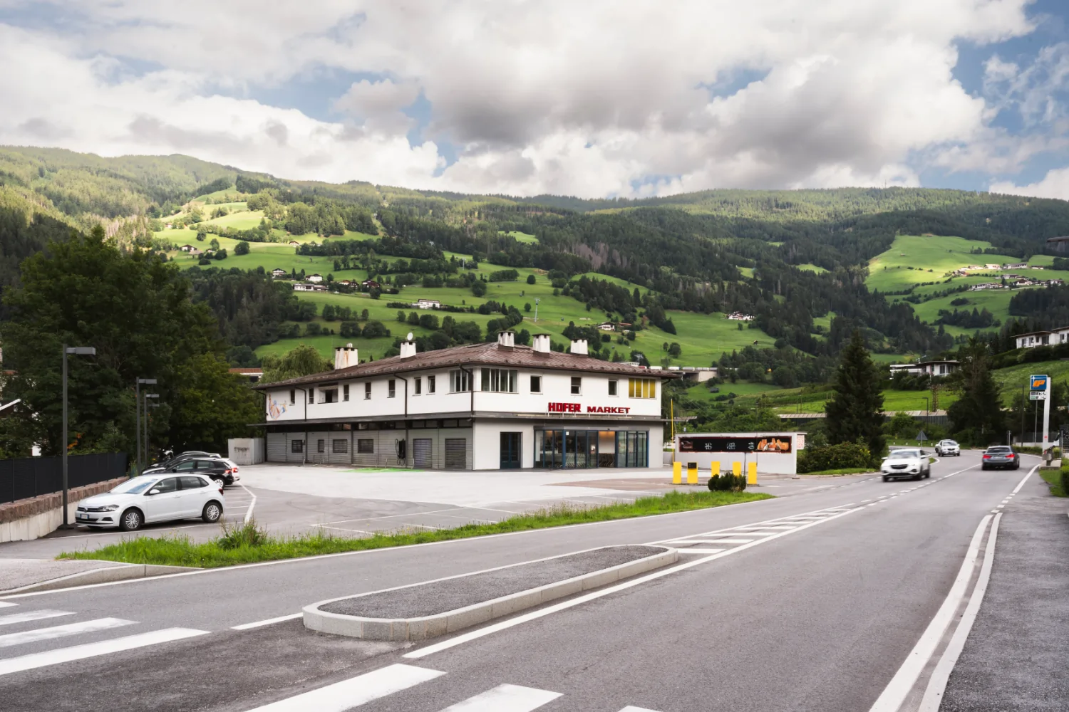 Der Hofer Market ist auf dem Bild neben einer Hauptstraße und einer alpinen Landschaft im Hintergrund zu sehen.